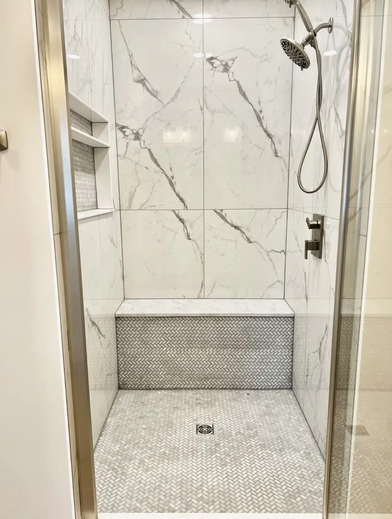 Newly remodeled walk-in shower with marble-look tile walls, built-in bench, herringbone mosaic floor, and brushed nickel fixtures — the type of bathroom plumbin