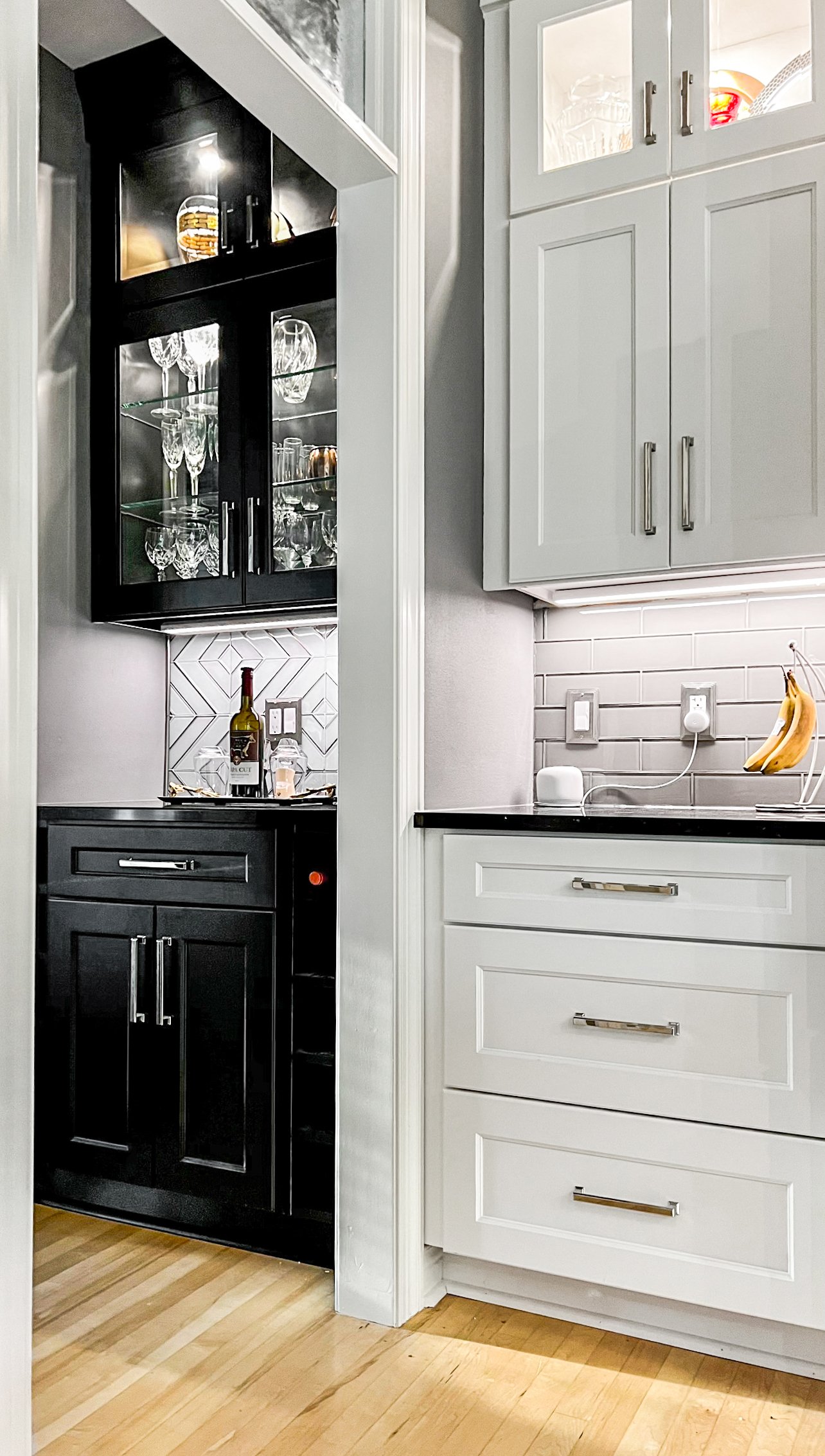 Two-tone kitchen remodel featuring light gray shaker cabinets with subway tile backsplash and black butler's pantry cabinetry with glass-front uppers, hardwood 
