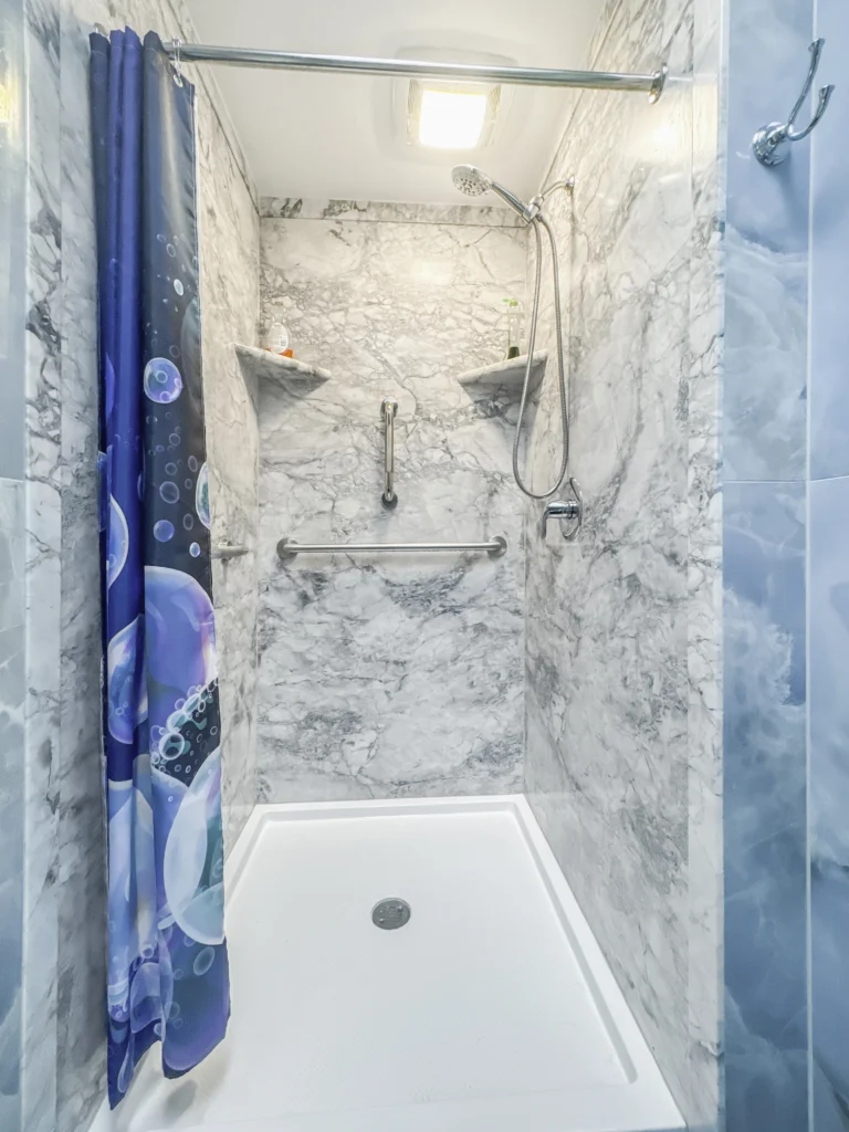 Finished walk-in shower with marble-look acrylic wall panels, white low-profile shower pan, chrome grab bars, handheld showerhead, built-in corner shelves, and