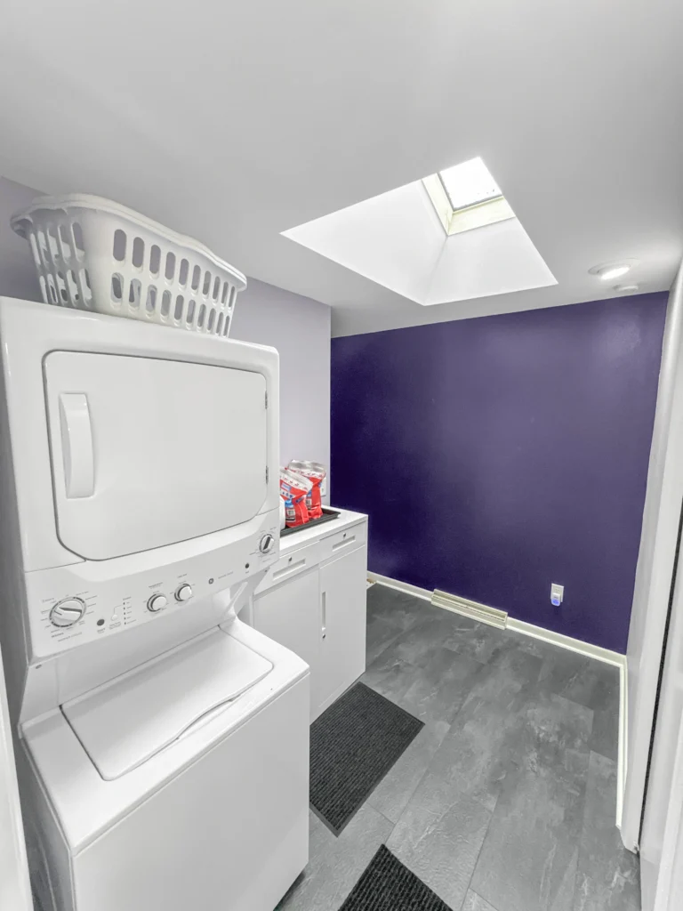 Compact laundry/mudroom with stacked GE washer-dryer, bold deep-purple accent wall, skylight, gray slate-look tile floor, and baseboard heater; laundry basket a