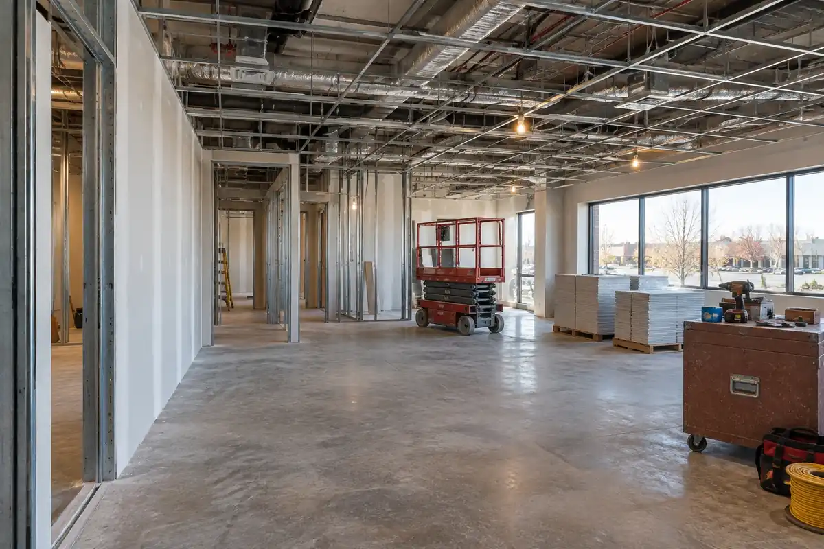 Leased office suite mid build out with metal stud partitions, drywall on one side, an open ceiling grid showing ductwork, a scissor lift and a window wall